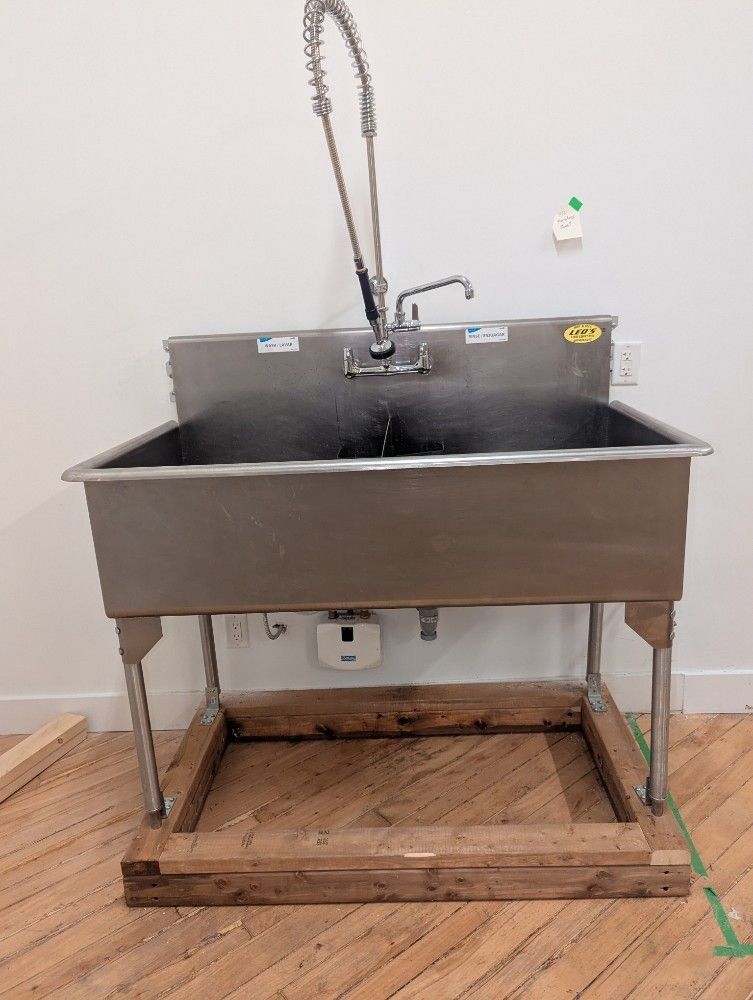 Commercial stainless steel sink with tall faucet, on wooden base in a room.