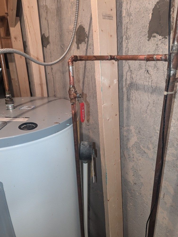 Water heater with copper pipes, a red valve, and wooden supports against a gray concrete wall.