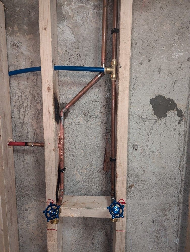 Copper plumbing pipes installed in a wooden frame against a concrete wall, with blue and red water shut-off valves.