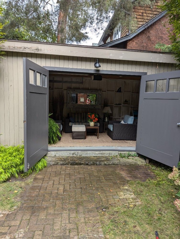 Open garage with gray doors, furnished as an outdoor living room with furniture, plants, and a mirror.