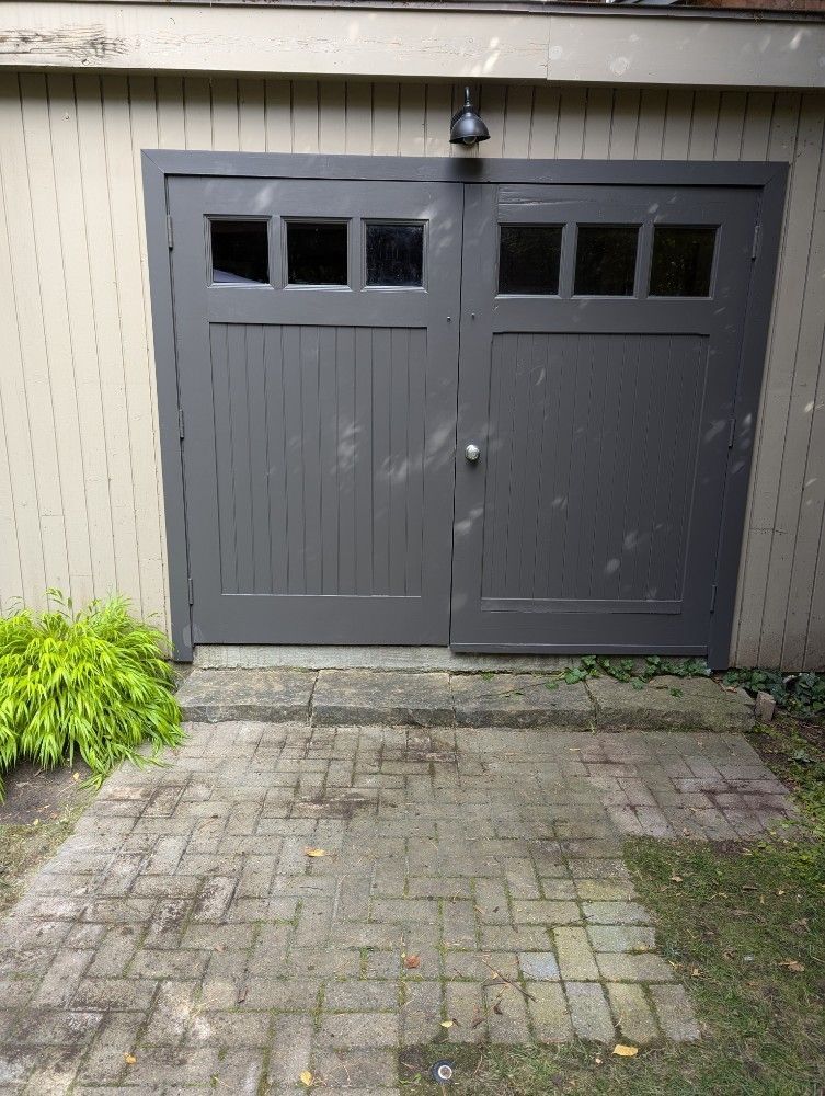 Gray double doors with window panes, brick walkway, and green foliage.