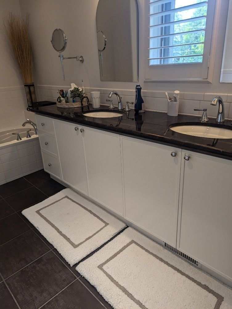 Bathroom with white vanity, black countertop, two sinks, white rugs, and window.