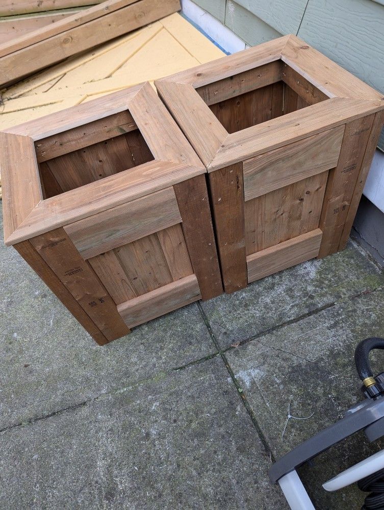 Two wooden planters side by side, outdoors on concrete, with a square opening and decorative trim.