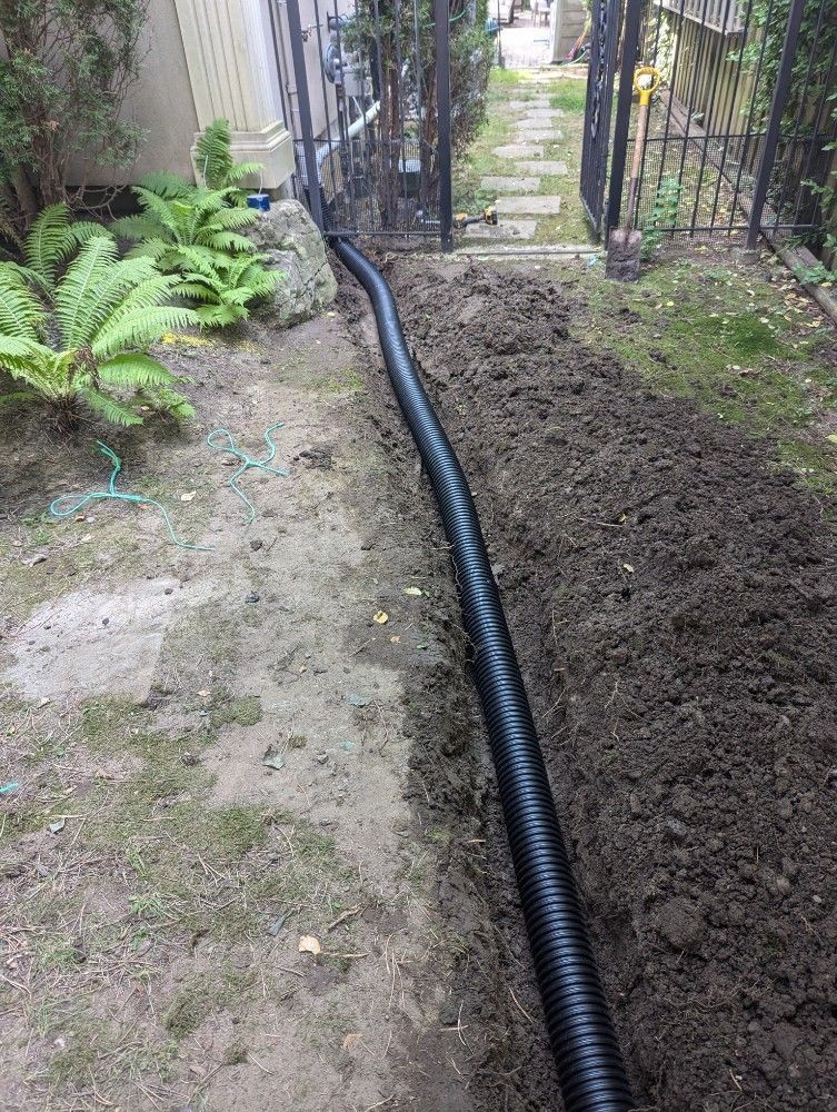 Black corrugated drain pipe in a trench beside a lawn, leading toward a gate in an outdoor setting.