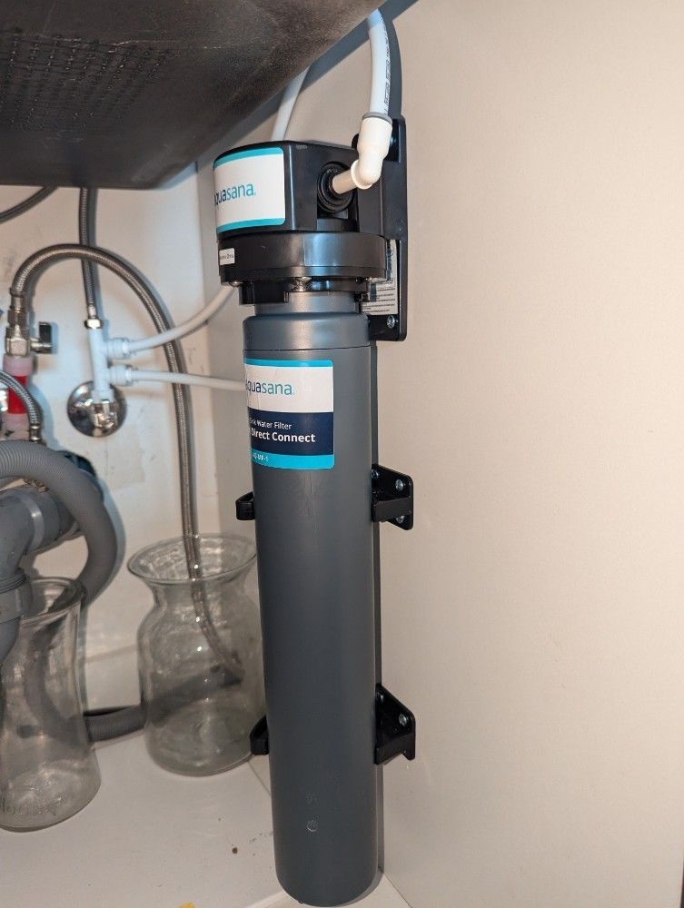 A water filter system installed under a sink. Gray filter housing, white tubing, and mounting brackets.