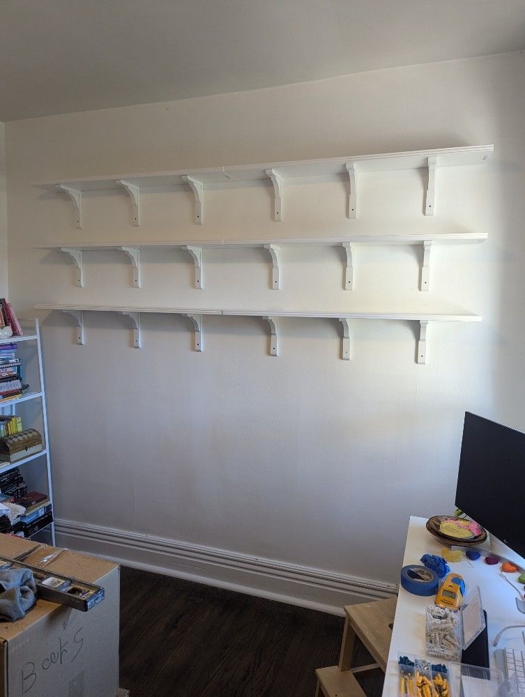 White shelves on a white wall, attached with white brackets. Room with dark floorboards and a desk.