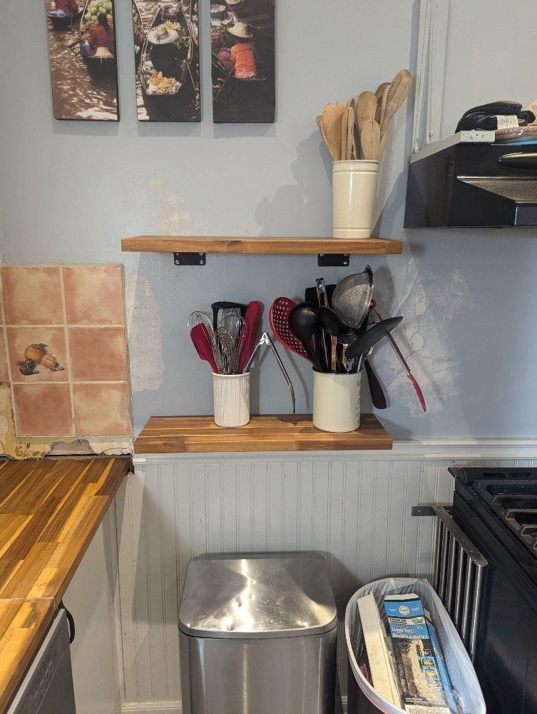 Shelves in a kitchen with utensils in ceramic containers, wooden countertops, and a stainless steel trash can.
