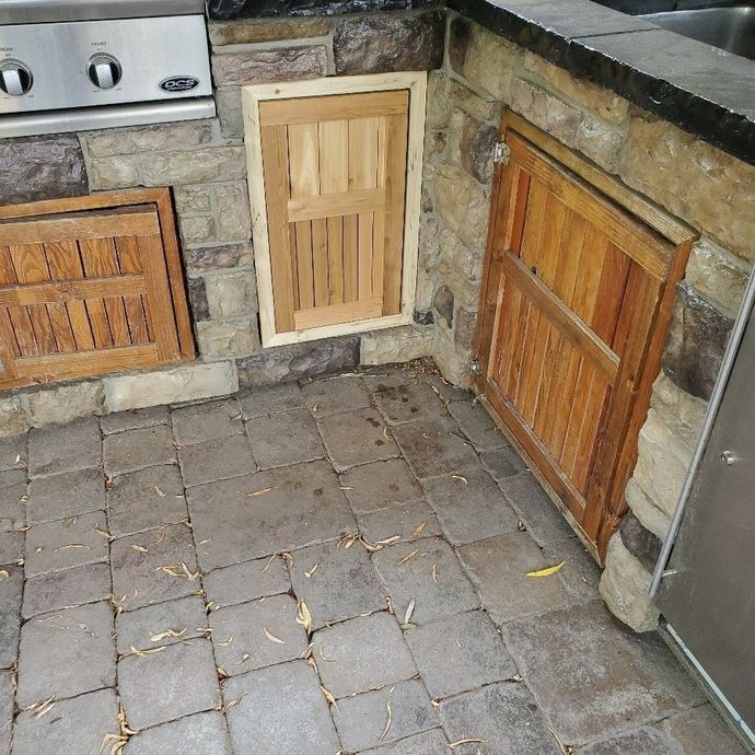 Outdoor kitchen with stone facade and wooden storage doors. Paved floor.