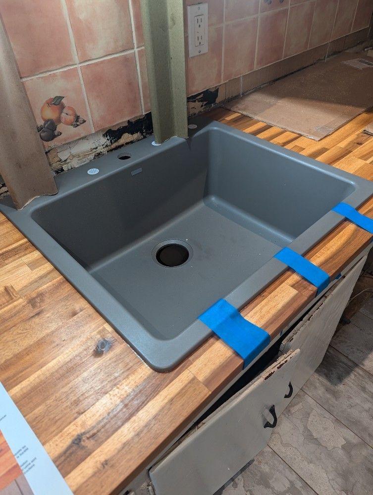 Gray kitchen sink installed in wooden countertop. Blue tape secures it. Cabinet below.