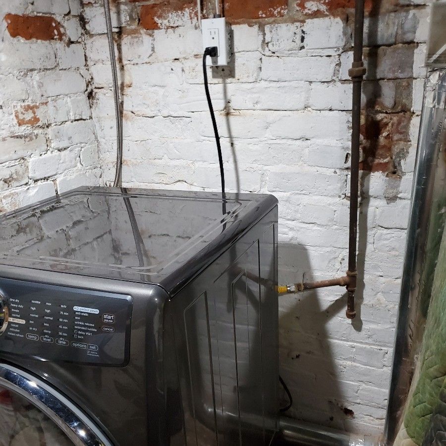 Washing machine in a basement with brick walls, electrical outlet, and copper pipes.