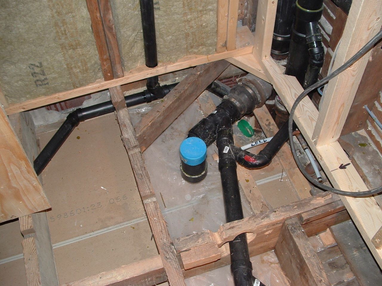 Plumbing pipes and connections inside a wall cavity with exposed wood framing. Black pipes, blue cap, gray surface.