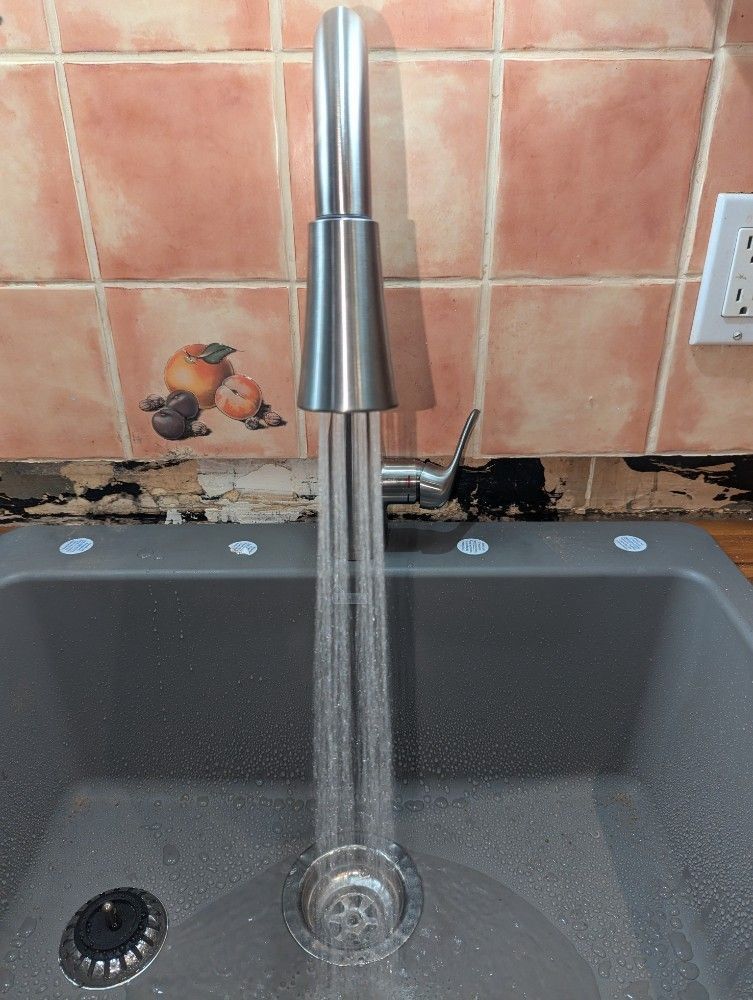 Faucet with running water over a gray sink. The backdrop is a tiled wall with some damage.