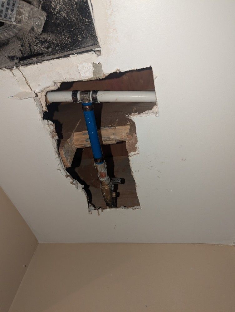 Hole in ceiling reveals water pipes. White, blue, and copper pipes visible in a damaged ceiling space.