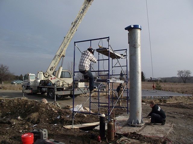 Construction workers near a tall metal well casing, using a crane and scaffolding in a field.