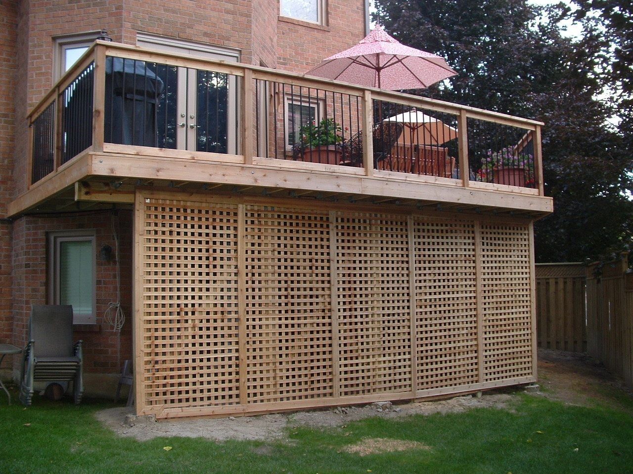 Wooden deck with trellis, attached to a brick house, small pink umbrella on deck, green grass below.