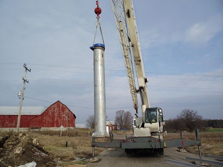 A crane lifting a tall, silver cylindrical object near a red barn under a cloudy sky.