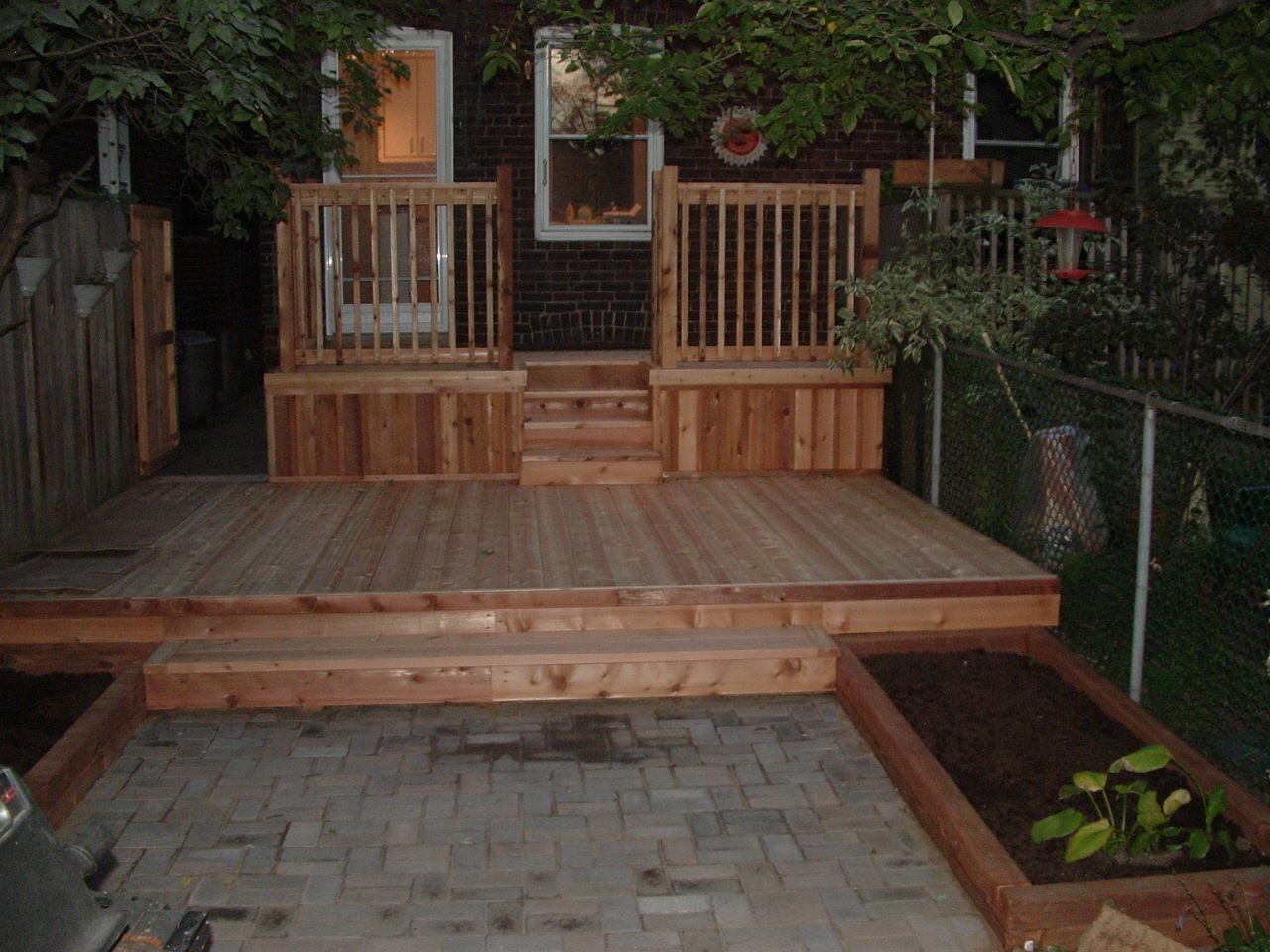 Wooden deck with steps, railings, and raised garden beds in a backyard, with a brick house in the background.