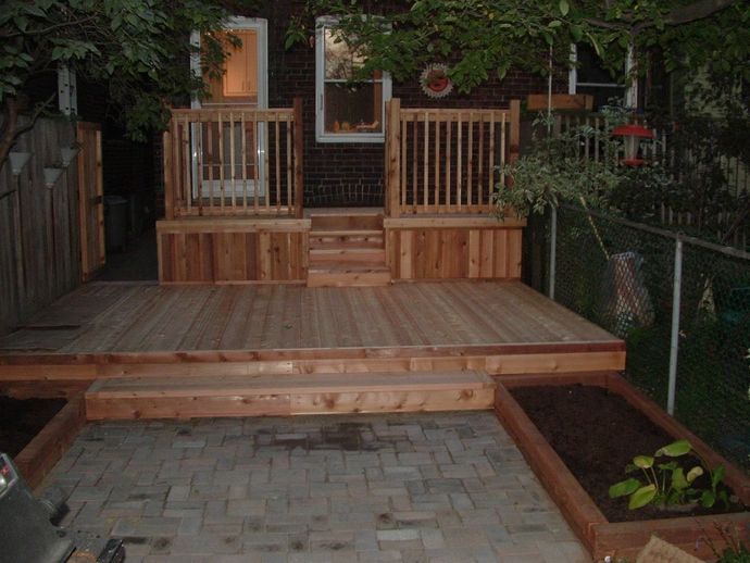 Wooden deck with steps, railings, and raised garden beds in a backyard, with a brick house in the background.