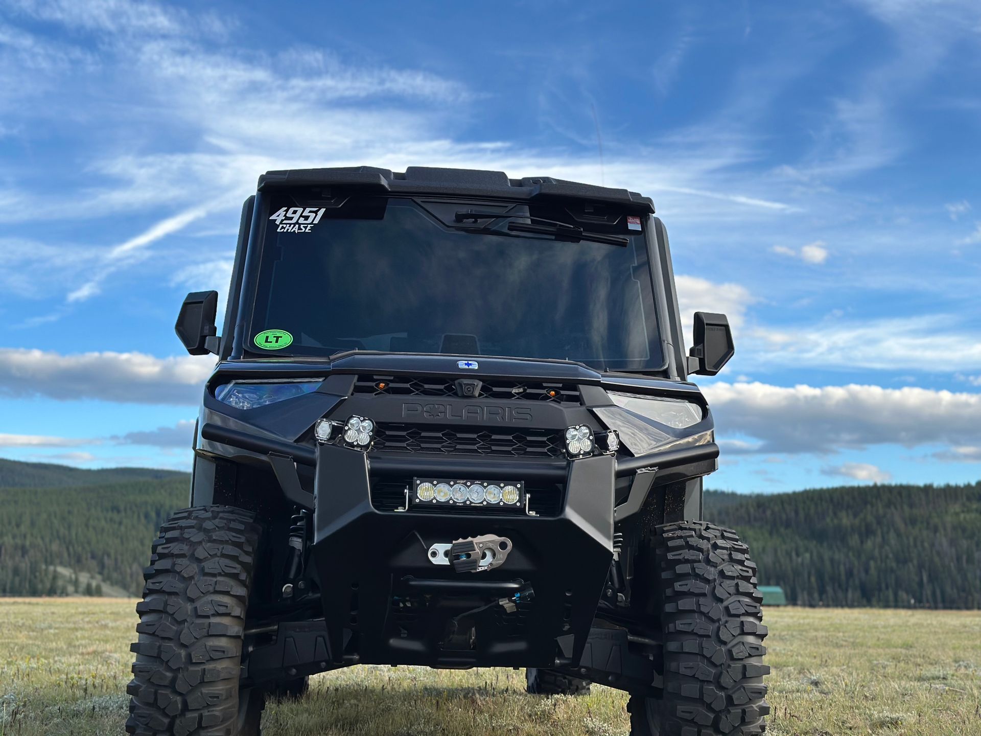 Black off-road truck with oversized tires parked in a grassy field under a blue sky.
