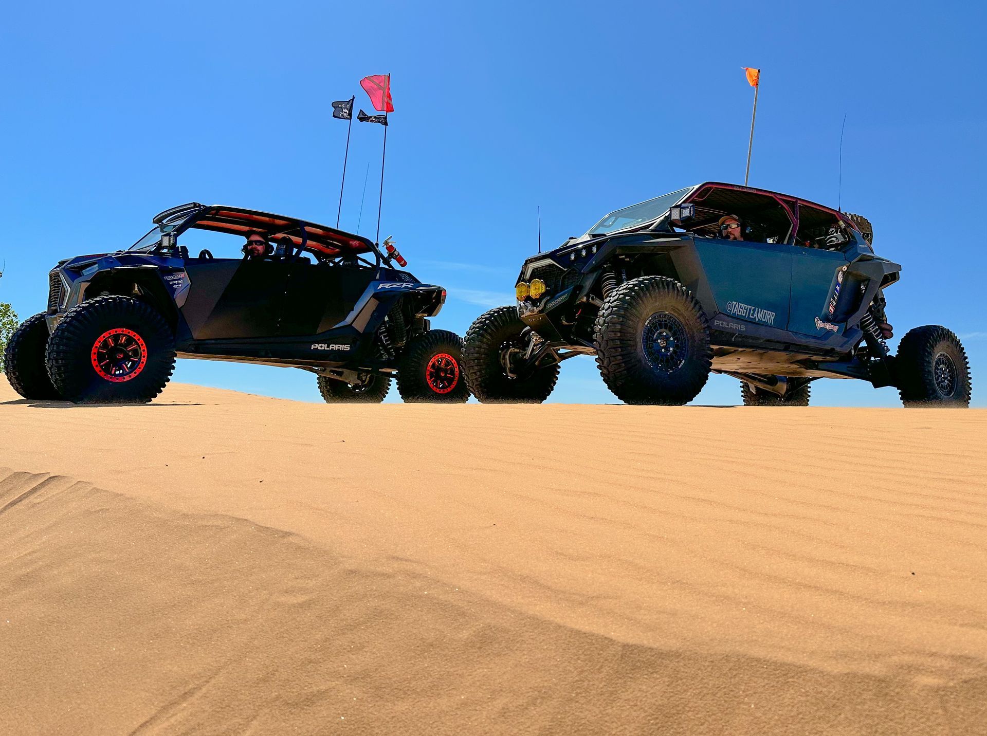 Two off-road vehicles with flags cresting a sand dune under a clear blue sky.