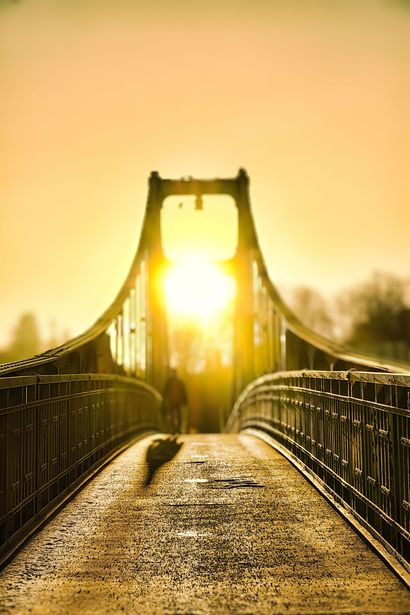 Ein sonnenbeschienener Hängebrückensteg mit Geländern und einem warmen, goldenen Sonnenaufgang vor uns.