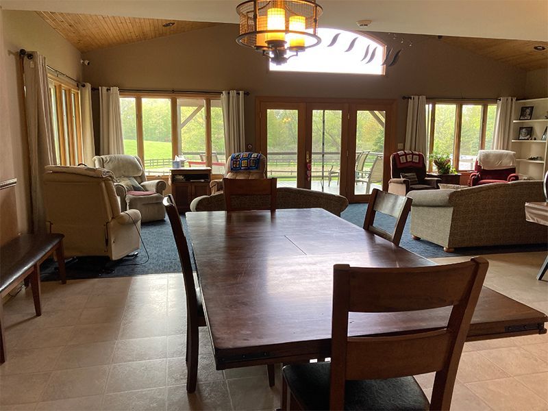 A dining room with a large wooden table and chairs, a living room with windows overlooking a green landscape.
