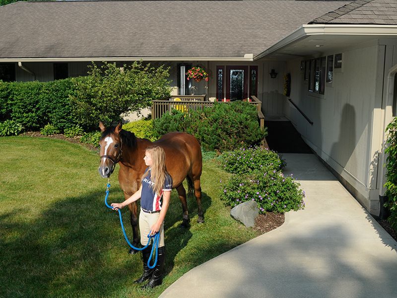 Woman in riding clothes leads a horse on a lawn in front of a house.
