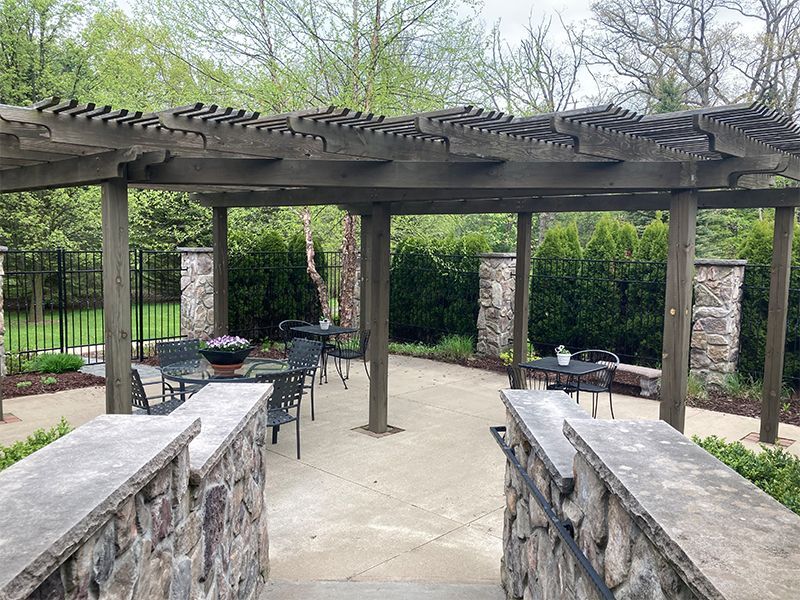 Stone patio with pergola, tables, and chairs. Green hedges border the patio, with steps in foreground.