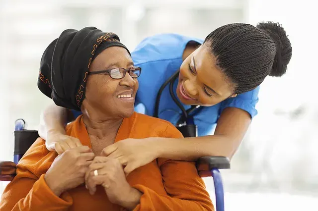 Nurse hugs and smiles at an older person in a wheelchair. Both are smiling.