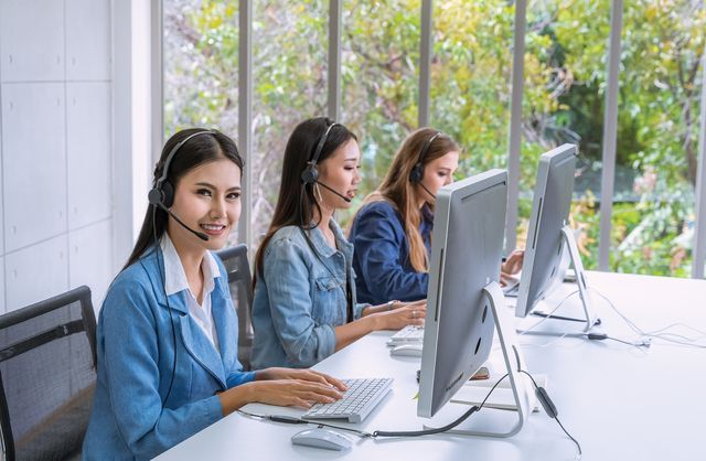 Three women wearing headsets are sitting at desks in front of computer monitors in a call center.