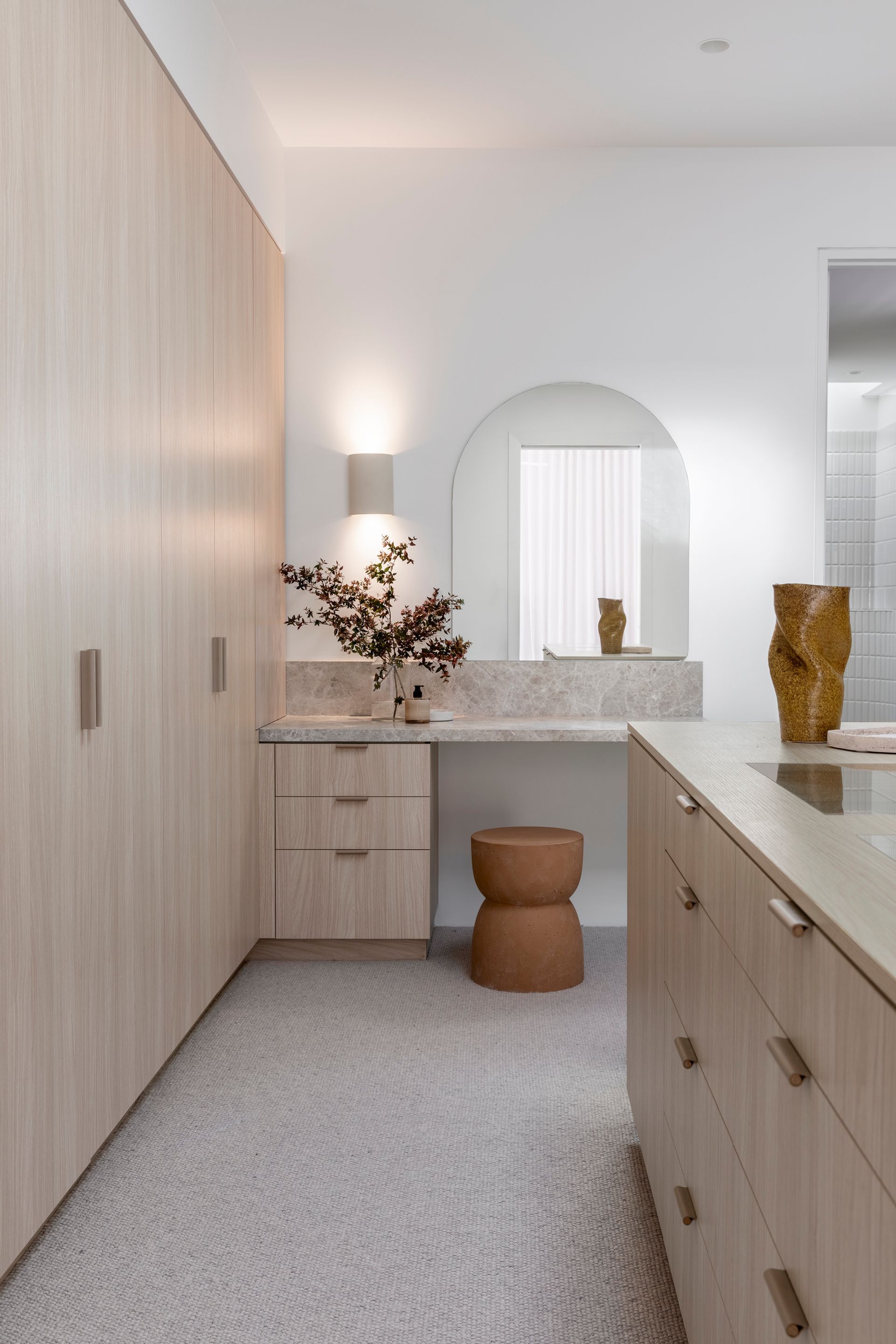 A neutral-toned dressing room with light wood cabinets, a vanity with an arch mirror, and a stool — Escarpment Building in Austinmer, NSW
