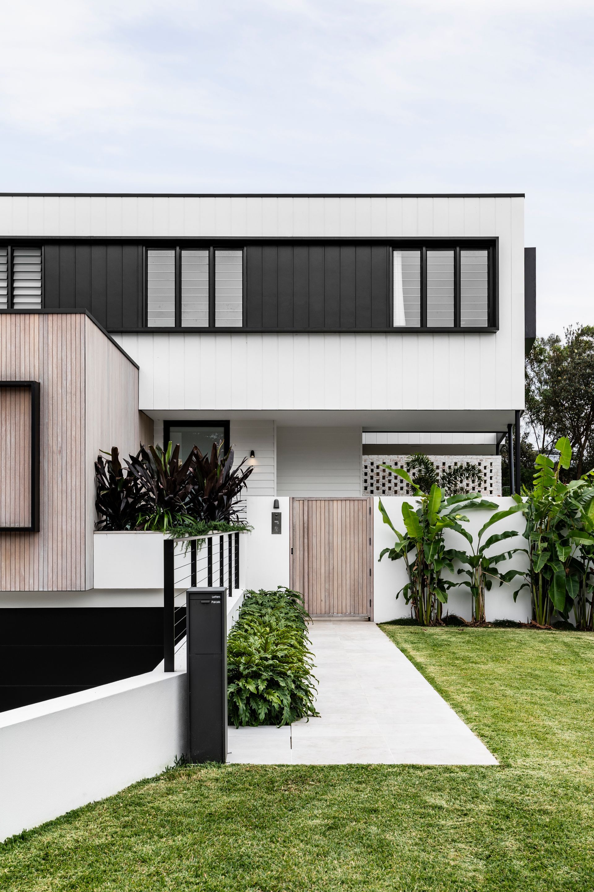 Modern two-story house with black and white facade, wood paneling, and a concrete pathway leading to a wooden gate — Escarpment Building in Austinmer, NSW