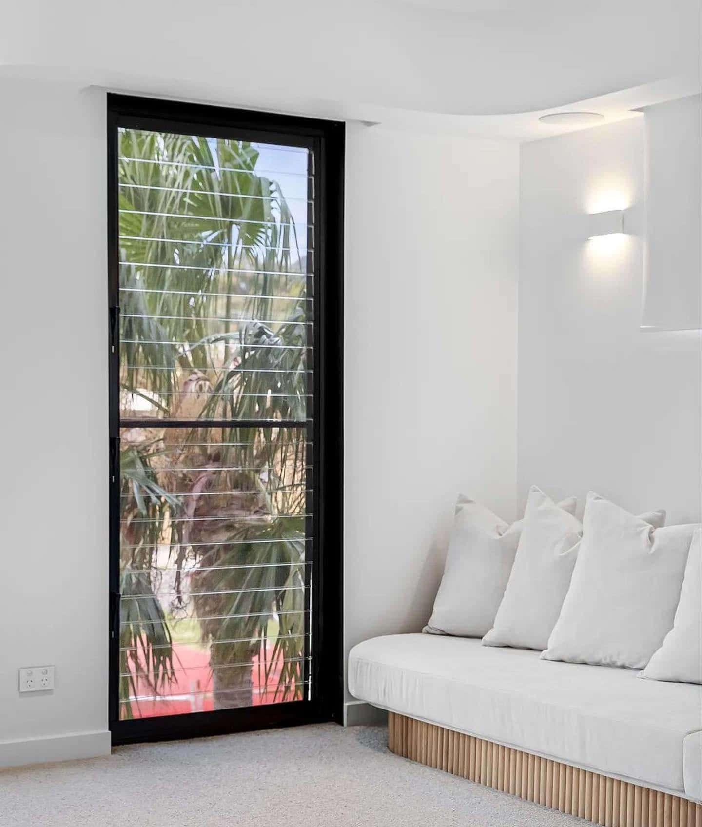 Black-framed glass door, white walls, and a white sofa with pillows. Palm tree visible through the glass  — Escarpment Building in Austinmer, NSW