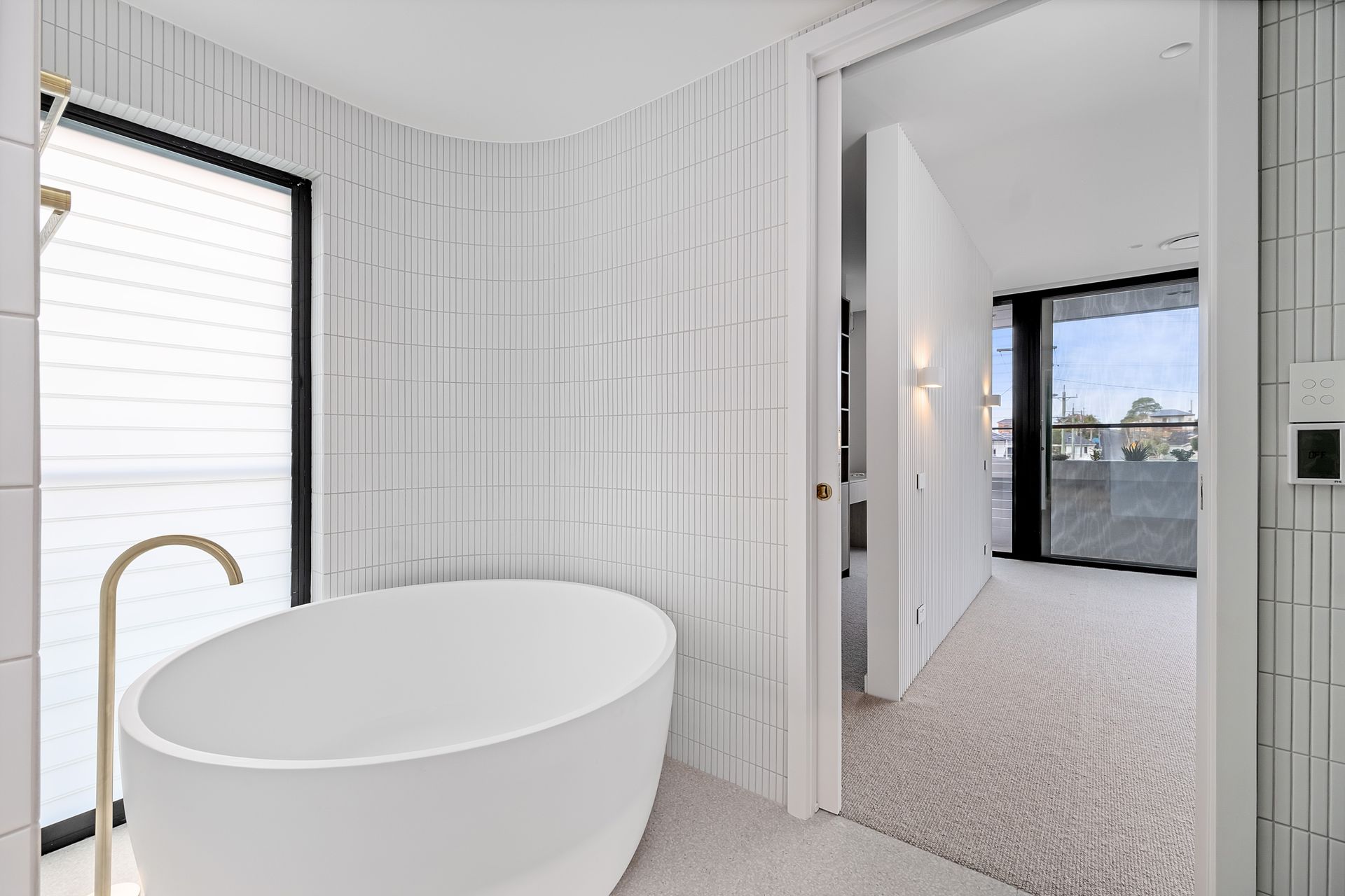 Modern bathroom with white tub, gold faucet, and textured white tile walls. View into hallway — Escarpment Building in Austinmer, NSW
