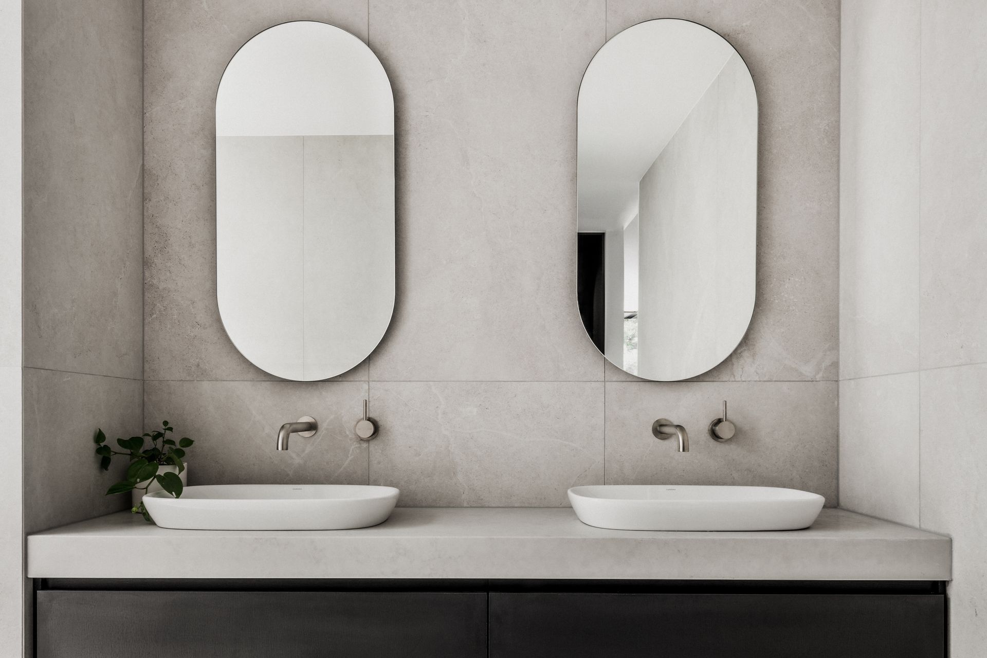 Bathroom with two oval mirrors above white sinks, gray tiled wall and concrete countertop — Escarpment Building in Austinmer, NSW