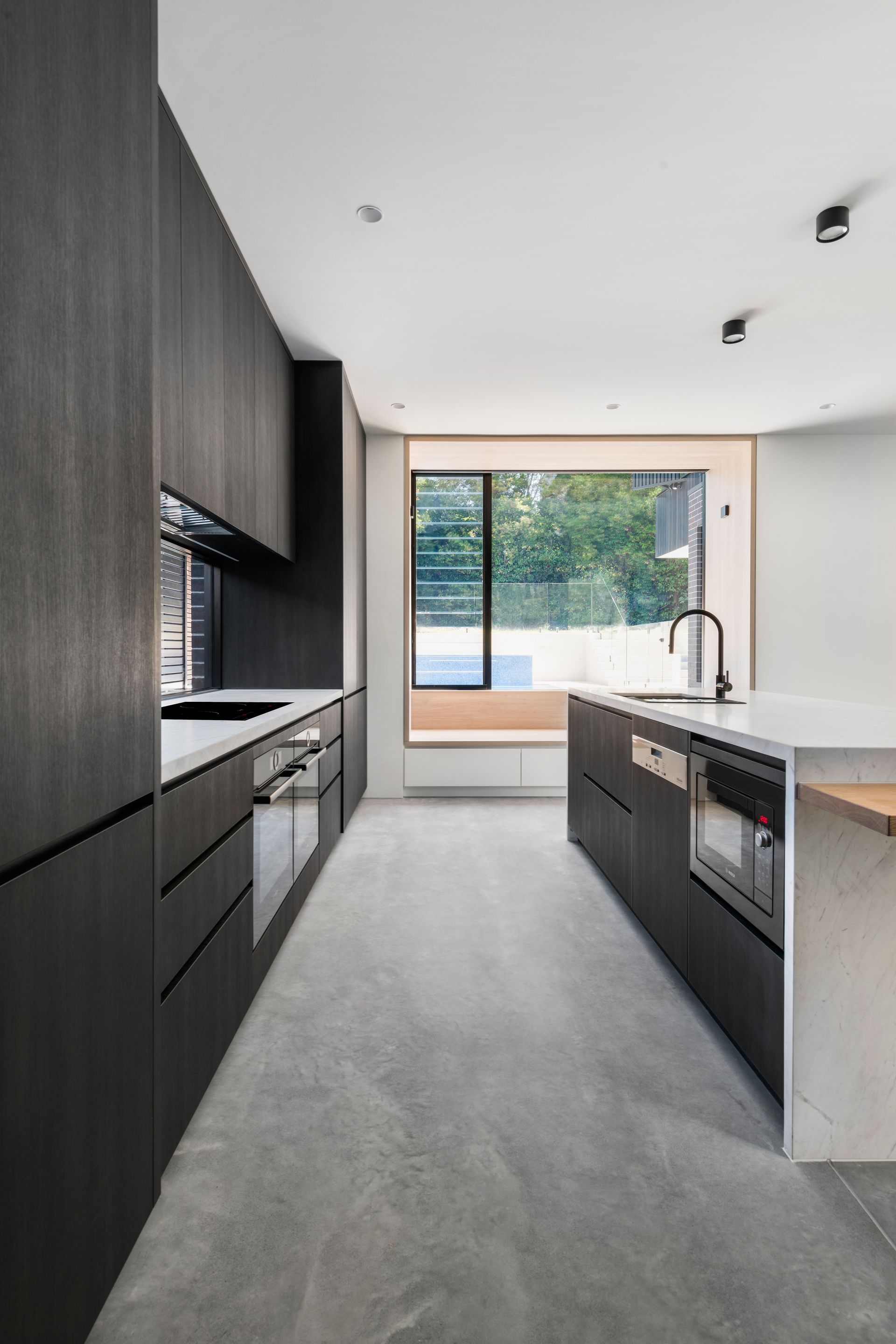Modern kitchen with dark wood cabinets, concrete floor, island, and large window — Escarpment Building in Austinmer, NSW