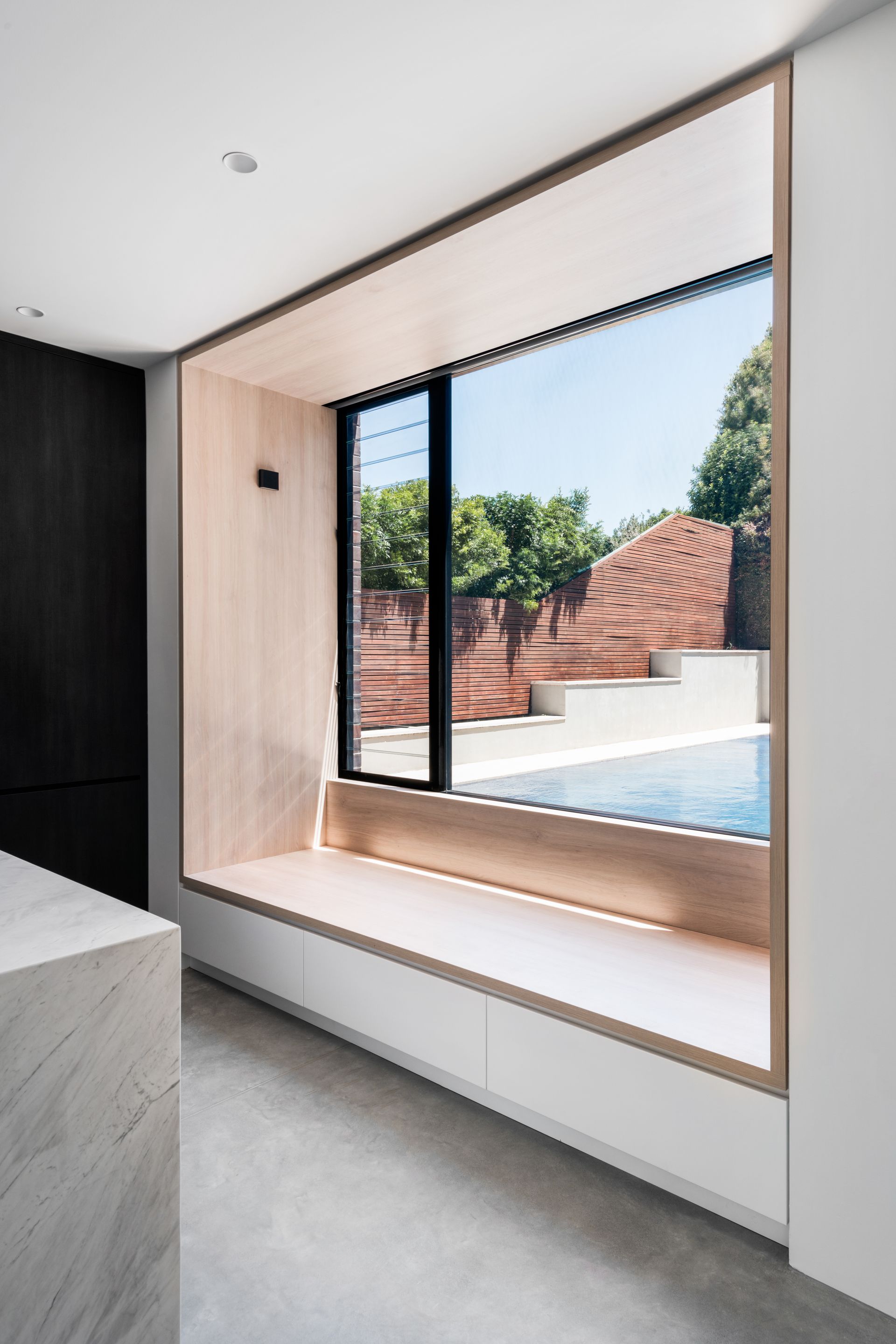 Window seat overlooking a pool. Light wood trim, black window frame, and white wall — Escarpment Building in Austinmer, NSW