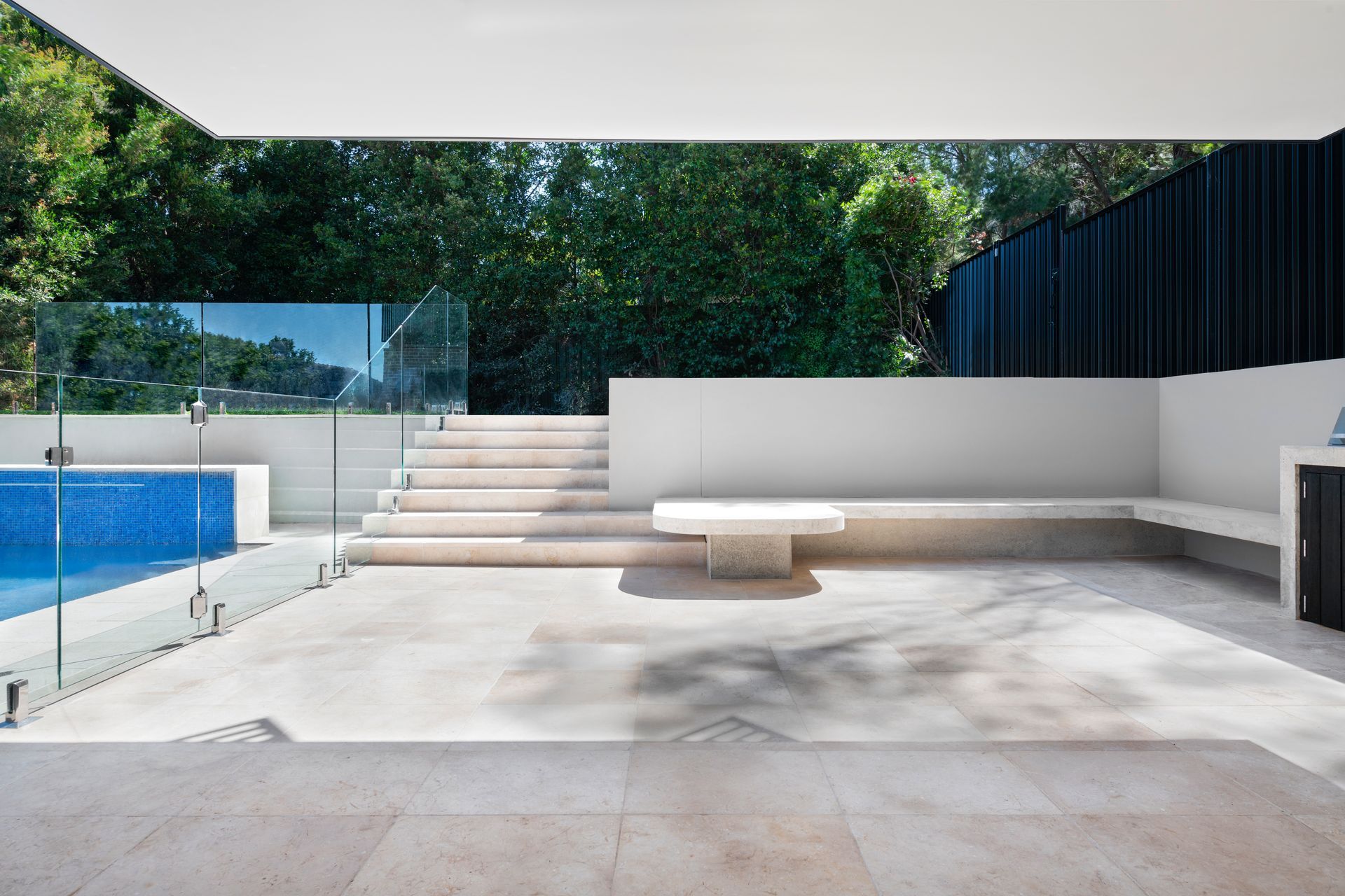 Patio with pool, steps, and built-in bench. Glass pool fence, light stone, and lush greenery in the background — Escarpment Building in Austinmer, NSW