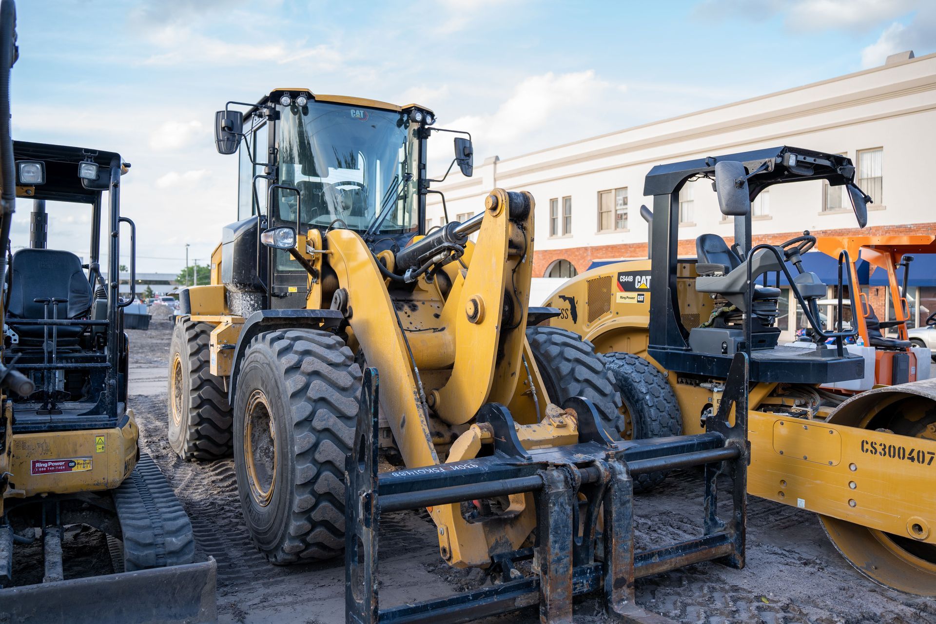 Yellow construction loaders parked in a gravel lot beside a building, with blue sky above