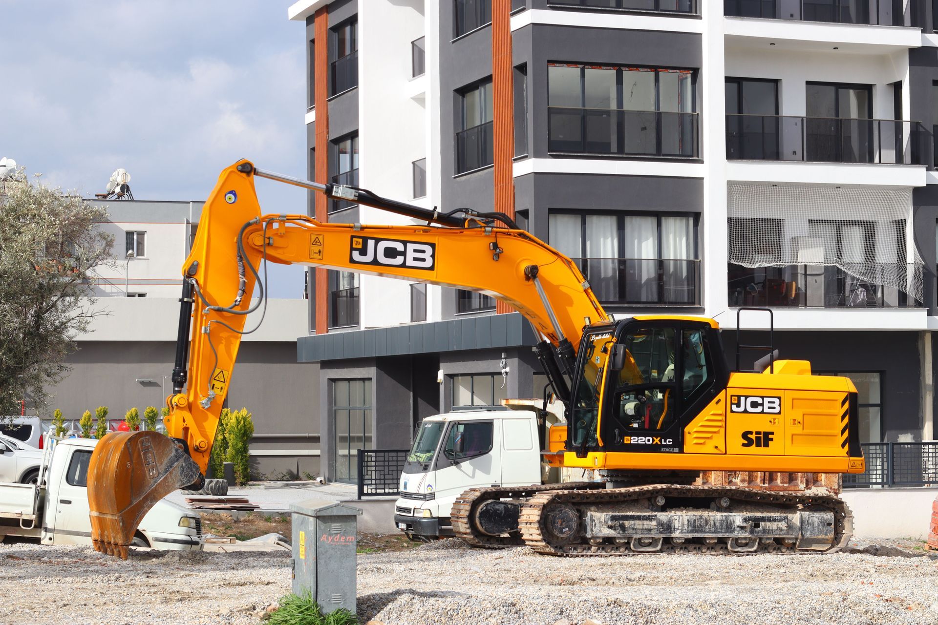 Yellow JCB excavator on a construction site beside a white truck and apartment building