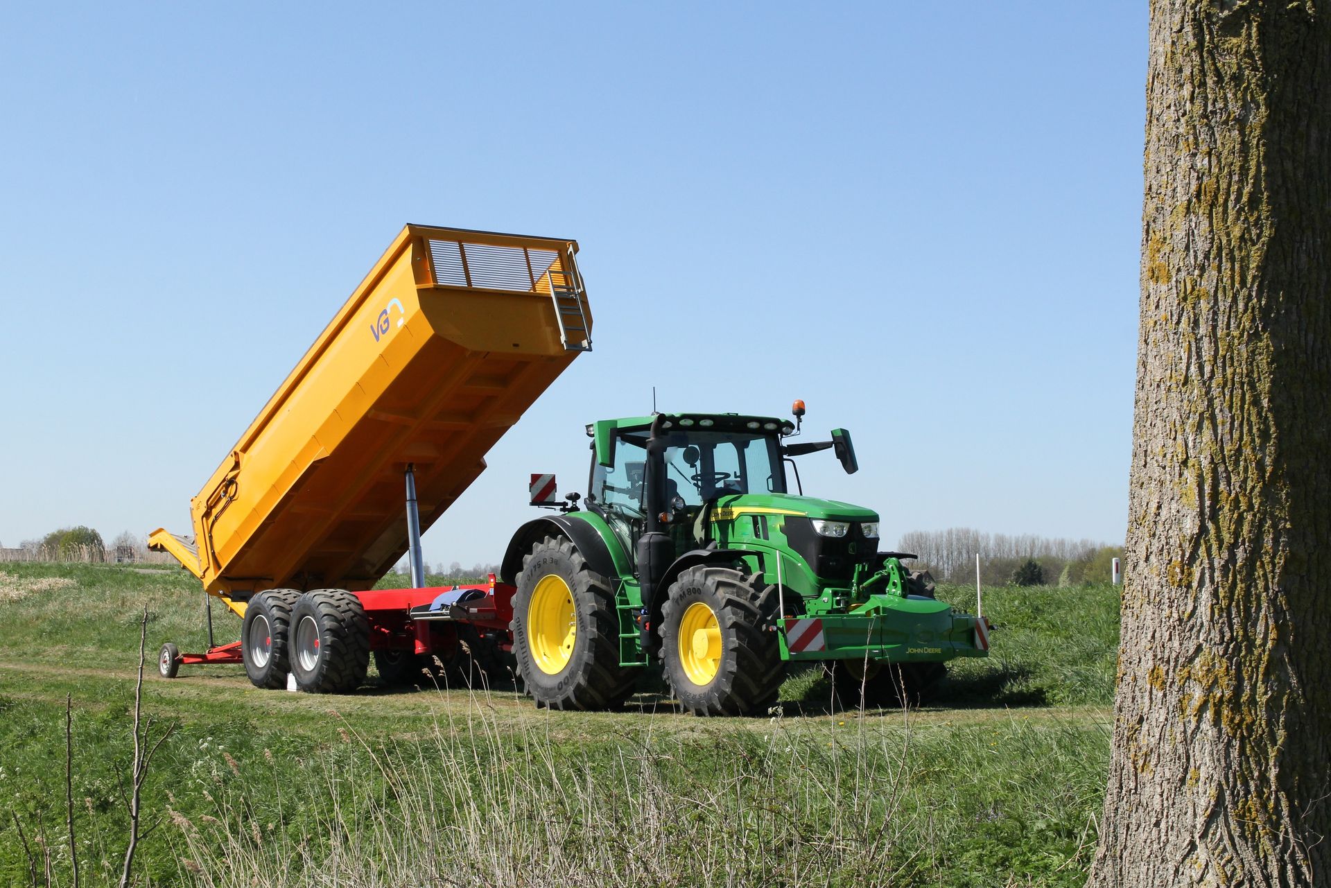 Green tractor unloading a raised orange trailer in a grassy field under a clear blue sky
