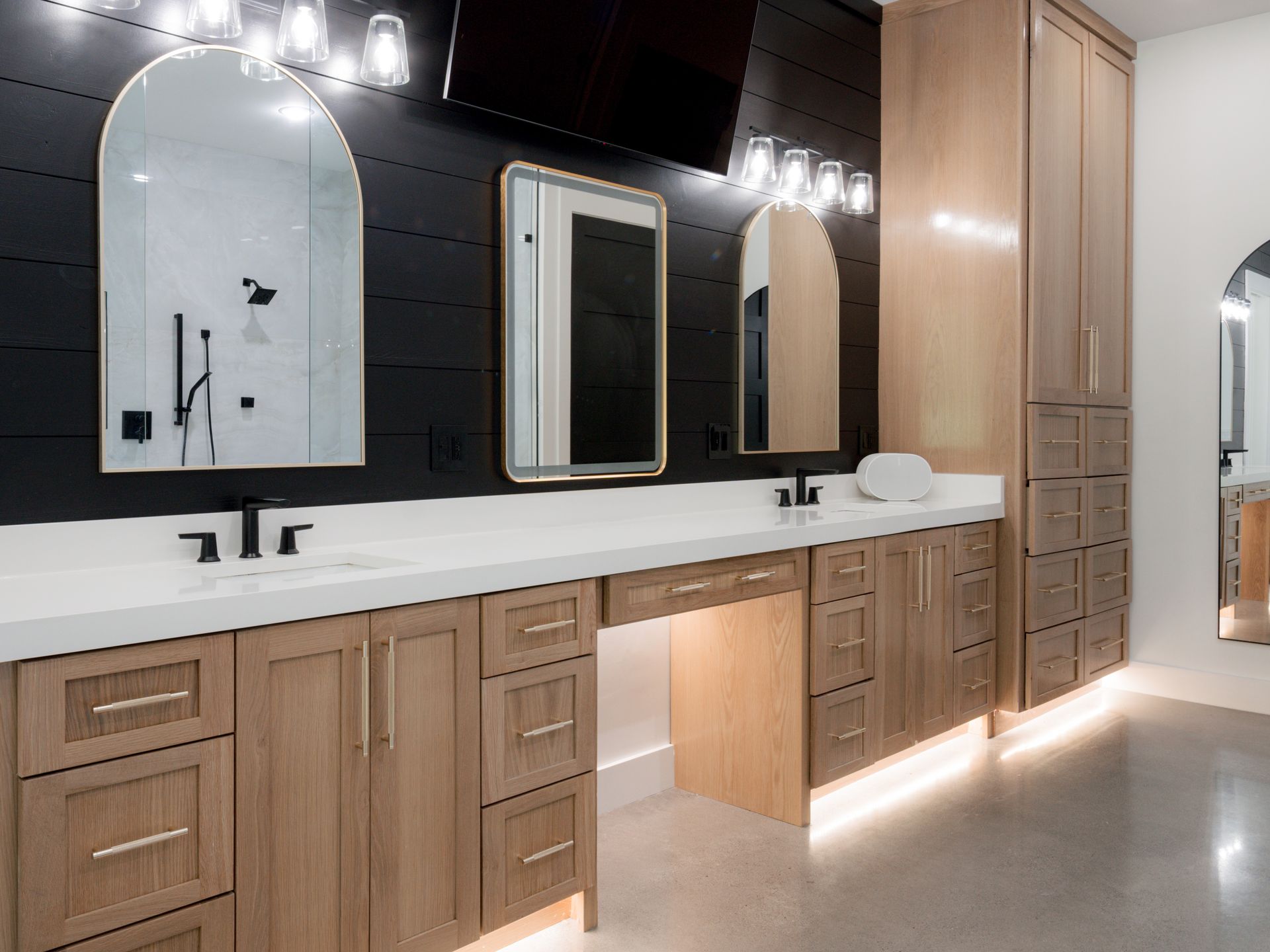 Modern bathroom vanity with wood cabinets, black wall, arched mirrors, and under-cabinet lighting