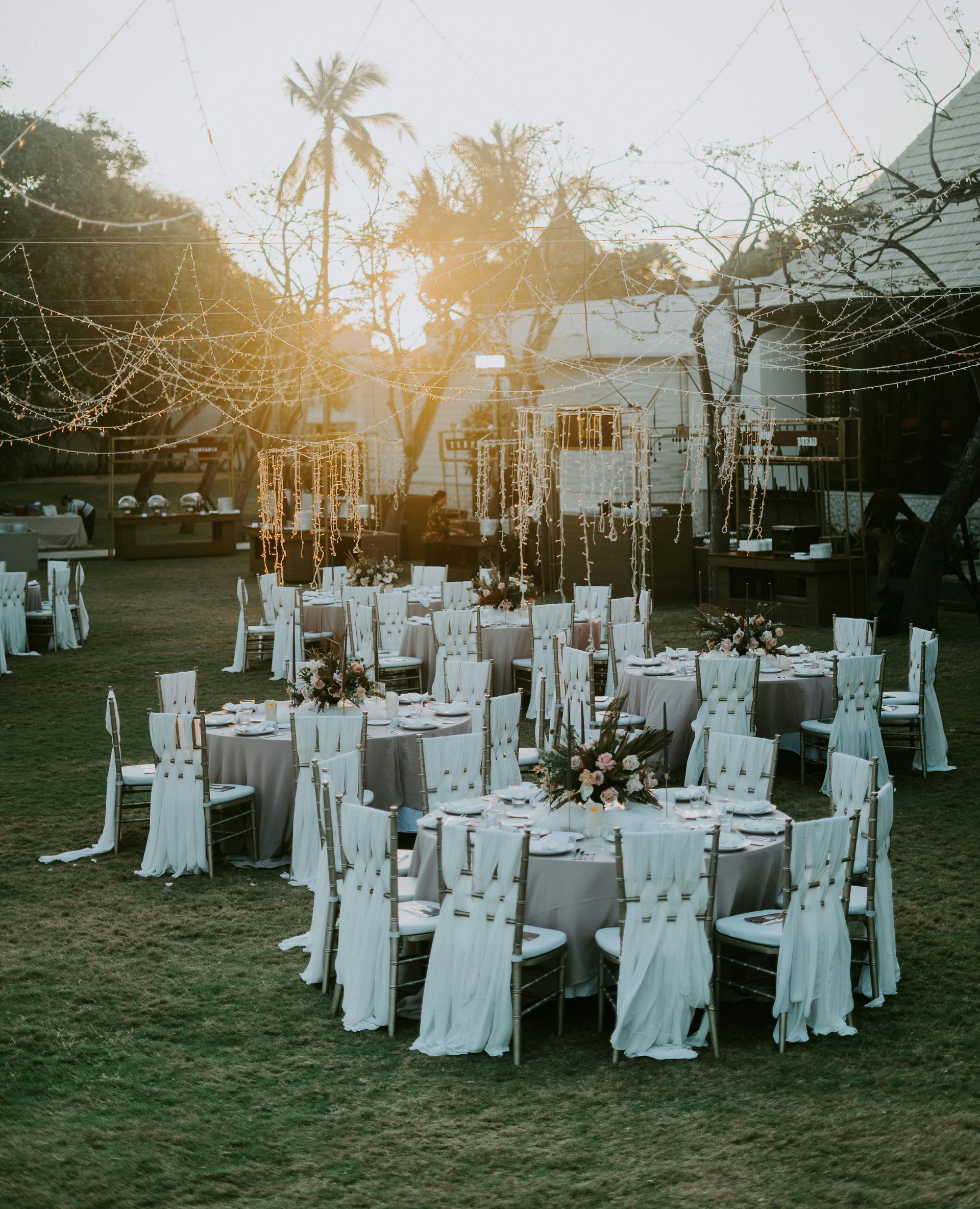 Outdoor wedding reception setup with round white tables and chairs on a lawn at sunset.