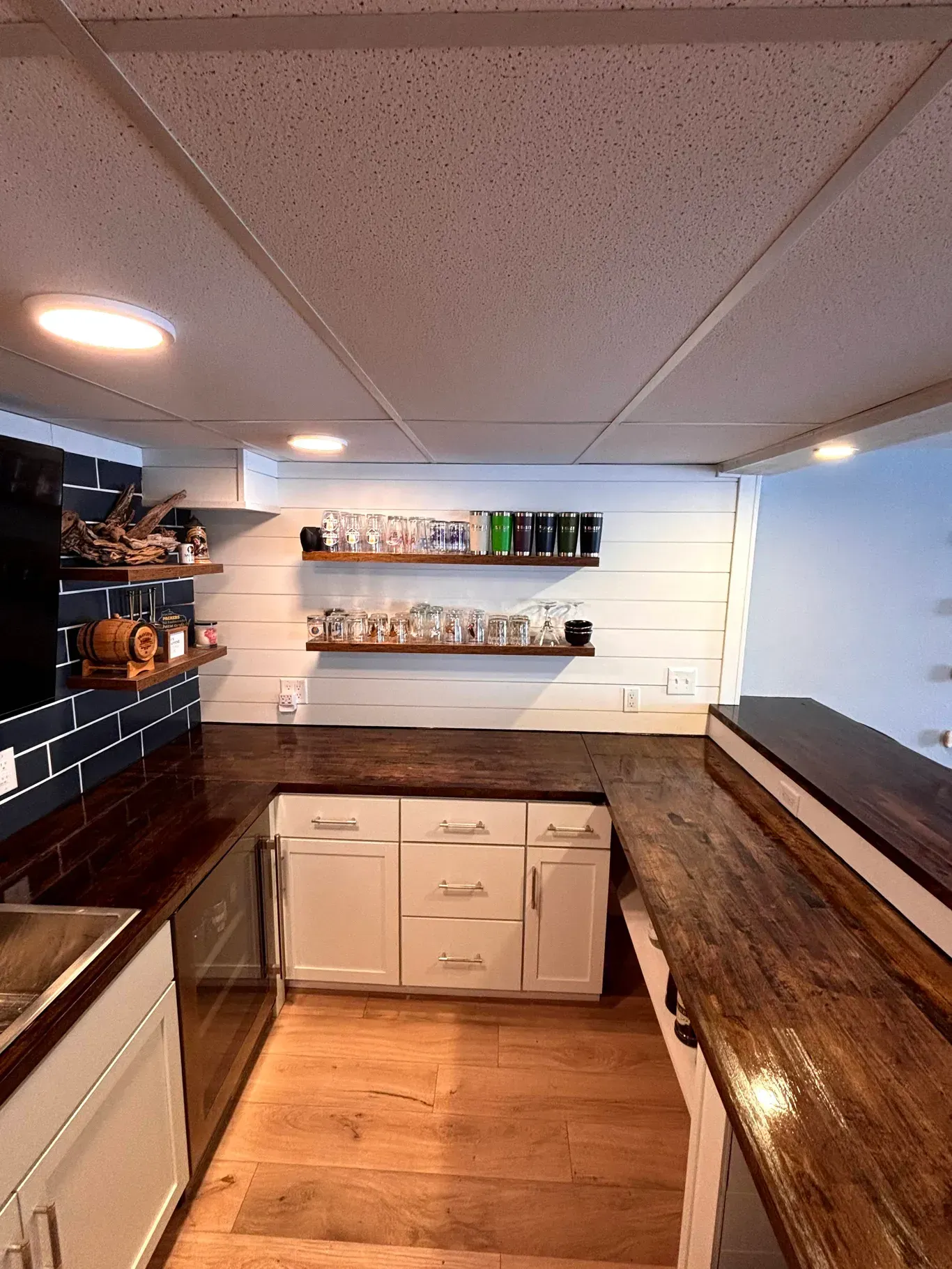 A bright kitchen with white cabinets, dark wood countertops, open shelving with glassware, and light wood-plank flooring.
