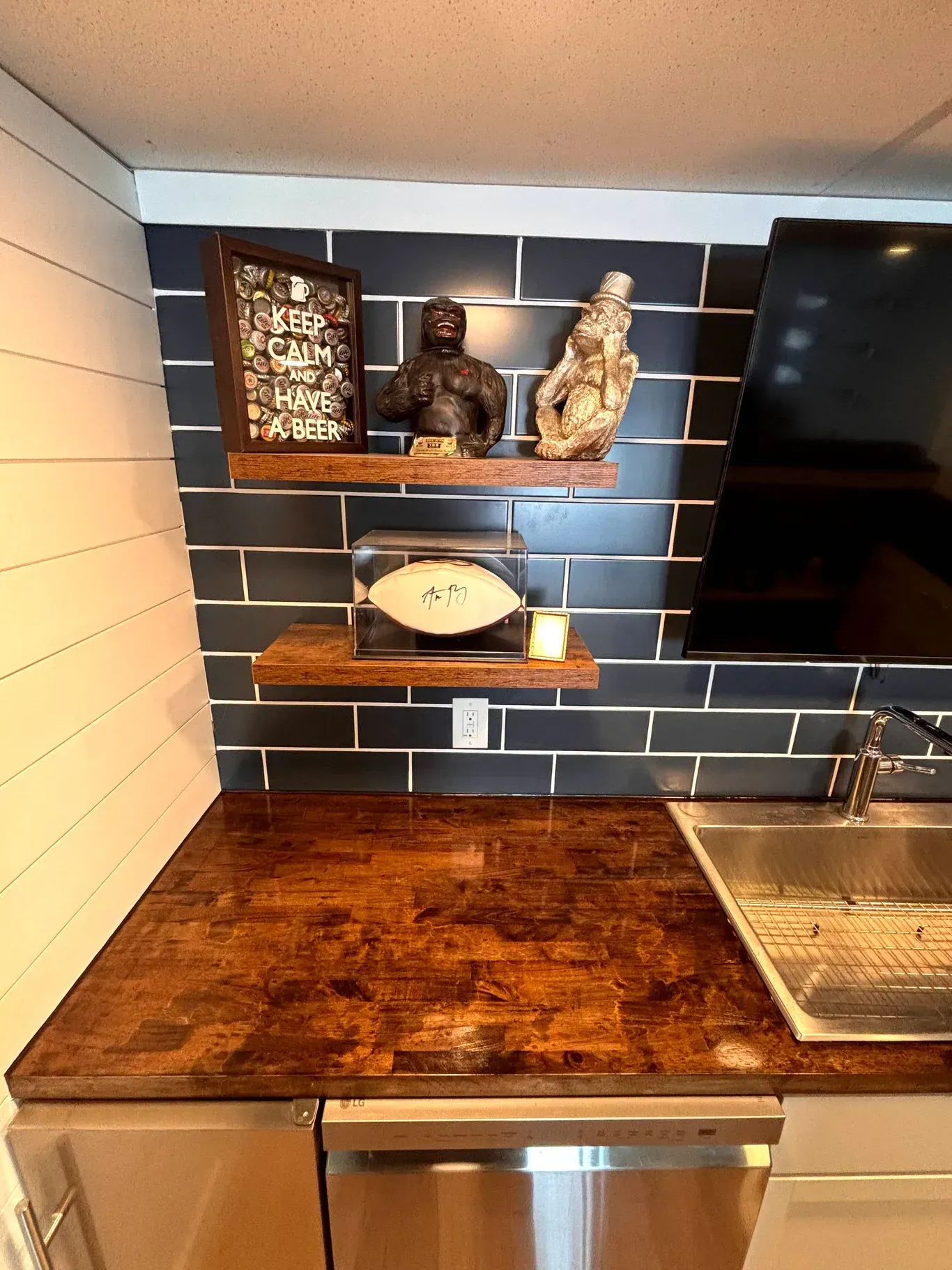 A dark-tiled kitchenette with wood countertops, two floating wood shelves holding decor, and a stainless steel sink.
