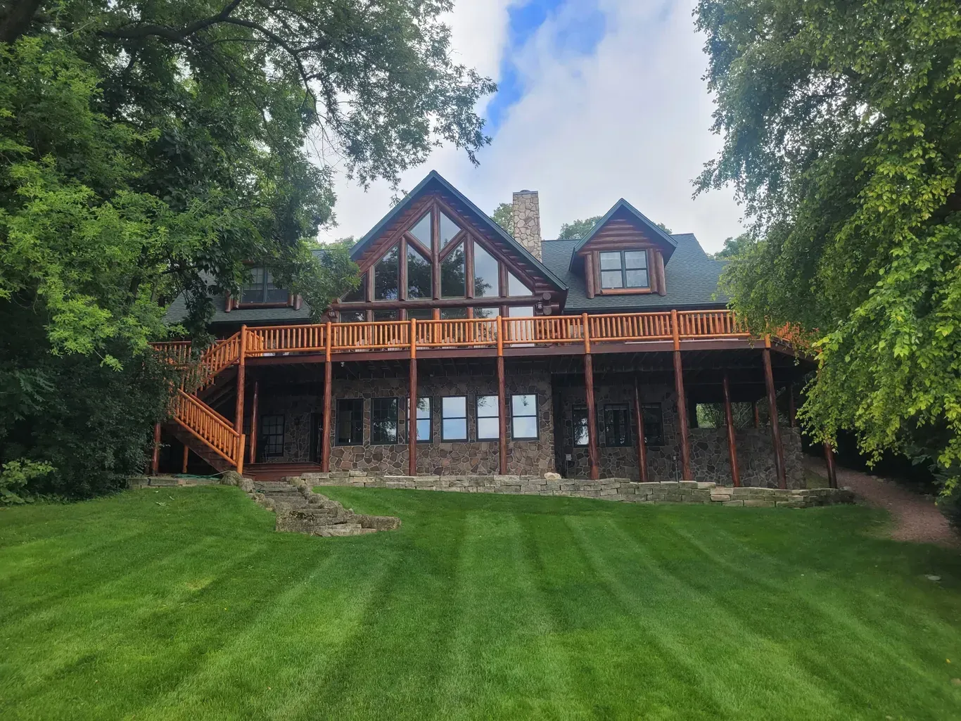 A multi-story wooden house with a stone base and large deck, surrounded by trees and a well-manicured lawn.