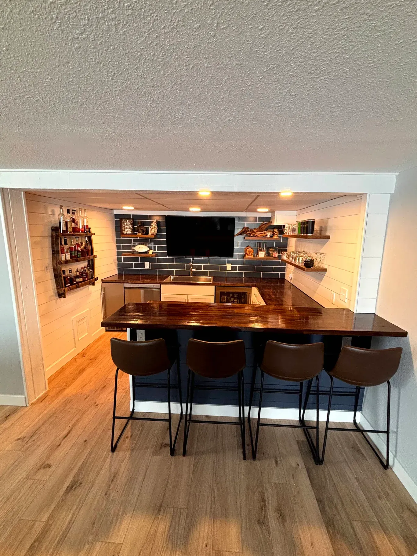 A home bar area featuring a dark wooden counter, four brown bar stools, white shiplap walls, and lit open shelving.