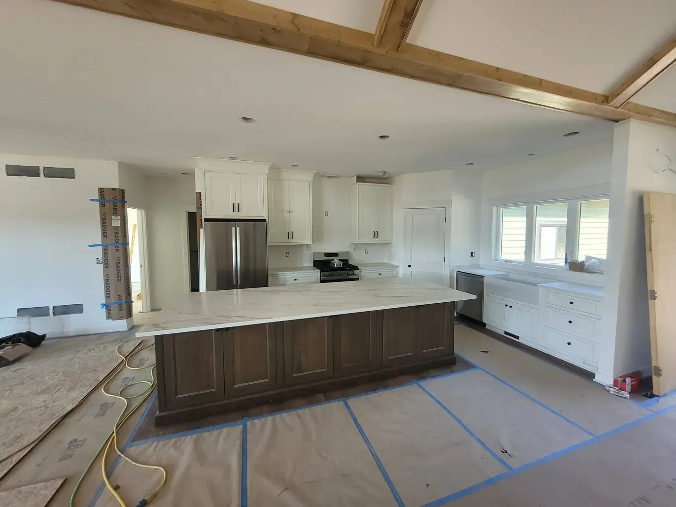 A kitchen under construction featuring a large dark wood island, white cabinets, stainless steel fridge, and exposed beams.