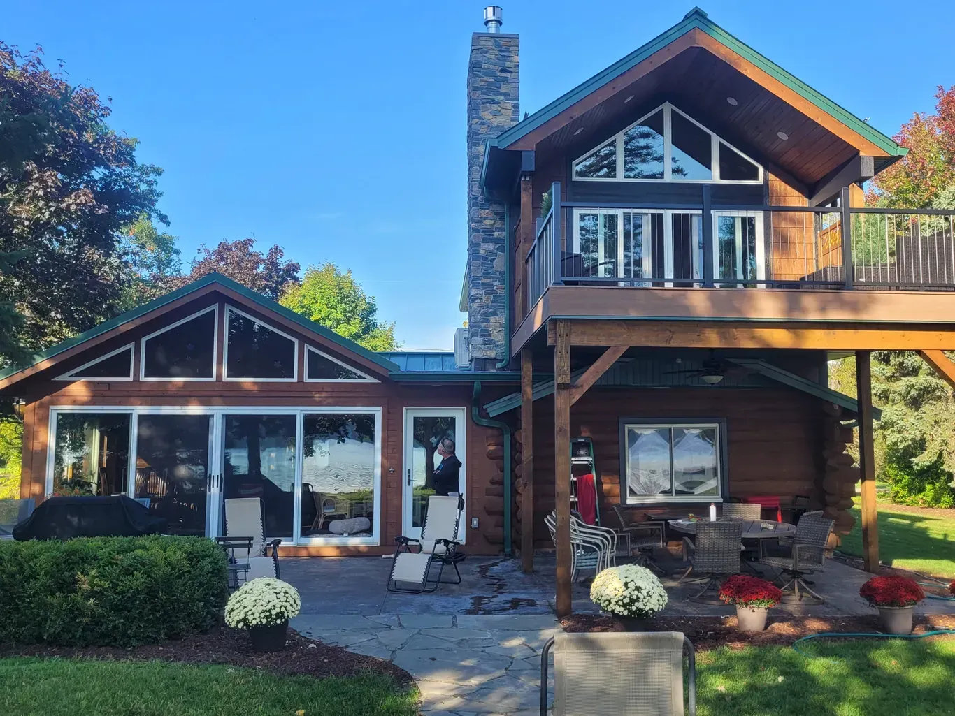 A two-story log cabin with a stone chimney, a glass-enclosed porch, and an upper deck, situated on a grassy lawn.