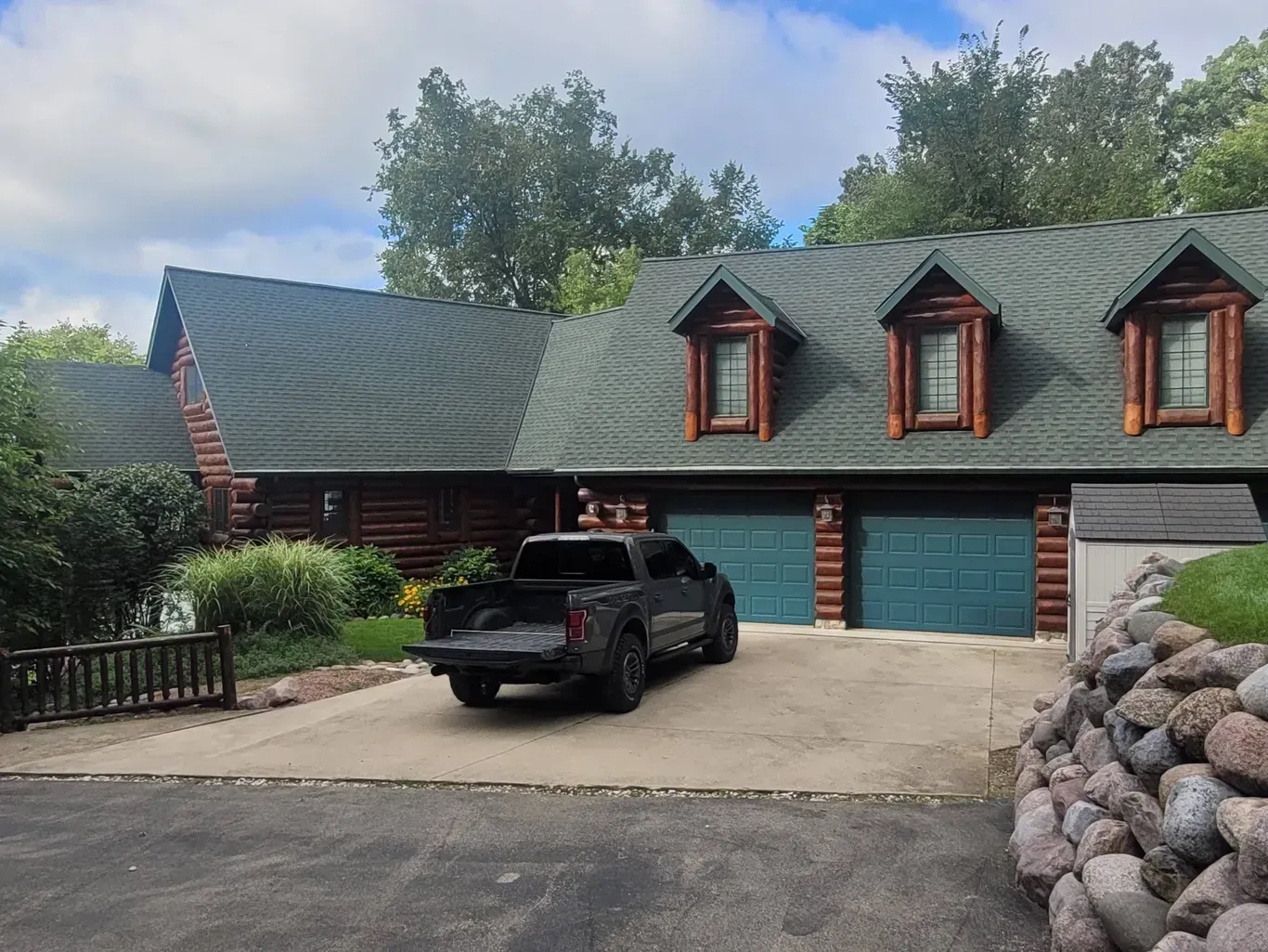 A log home with a green shingled roof, three gabled dormers, two garage doors, and a pickup truck parked in the driveway.